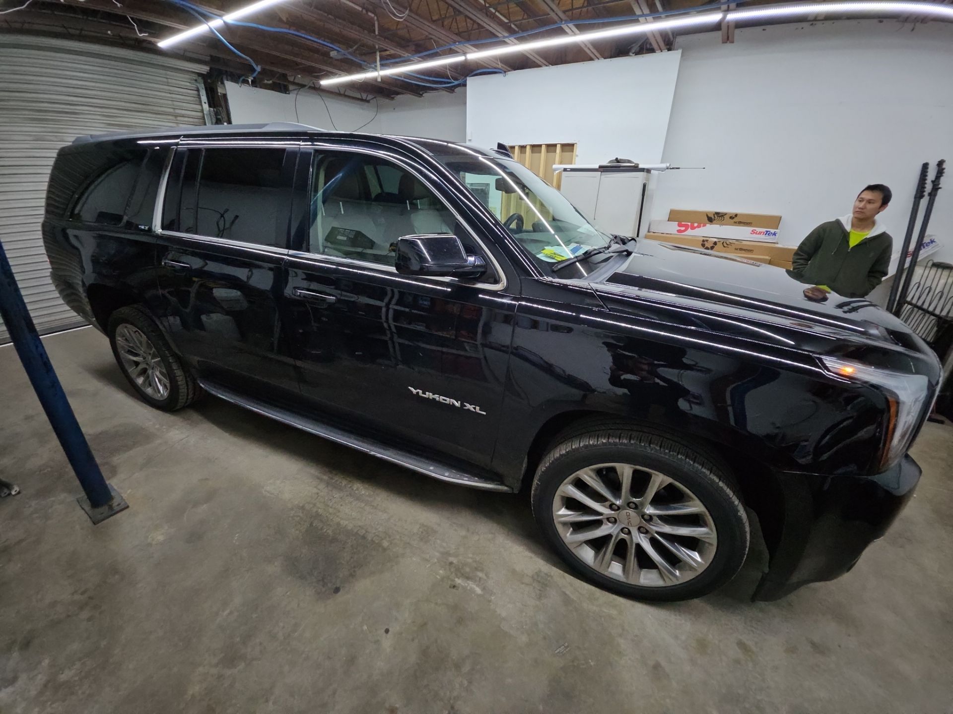 the image shows a black suv which is likely receiving a window tinting service the vehicle appears to be a yukon xl suggesting it is large and spacious the technician is present indicating that they are in the process of applying tint to the windows this service typically aims to enhance privacy reduce heat and protect the interior from uv damage the workspace appears organized and suitable for professional tinting