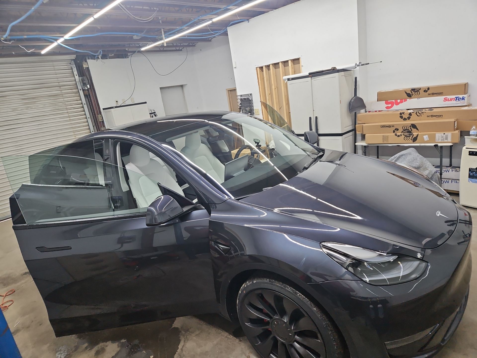 the image shows a tesla model y being serviced for window tinting the car has its side doors open indicating that the windows are being prepared for the application of tint the interior appears clean with white seats and the workspace is organized the tinting process is likely taking place to enhance privacy reduce glare and protect the interior from sun damage