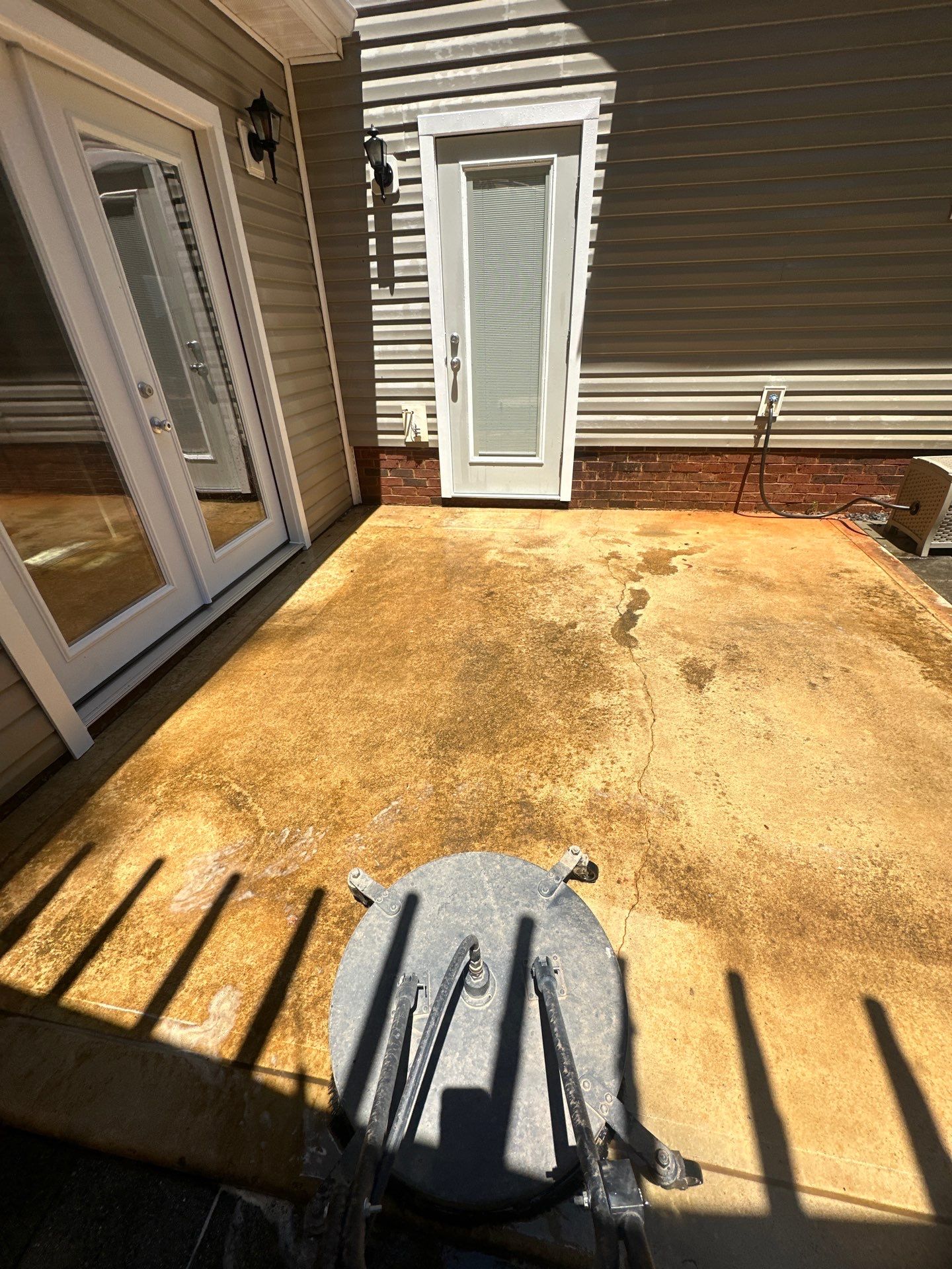 the service shown in the photo is pressure washing a concrete patio area this method uses high-pressure water spray to remove dirt grime mold and other debris from outdoor surfaces the pressure washing equipment visible in the image includes a surface cleaner attachment which is a round disk-shaped tool with nozzles underneath that evenly clean flat surfaces the difference in the color of the concrete where the surface has been cleaned compared to the dirty area is quite distinct indicating the effectiveness of the pressure washing process the cleaned area is much lighter while the area still to be cleaned shows discoloration and staining this is a common service offered by pressure washing companies to help maintain and restore the appearance of outdoor surfaces