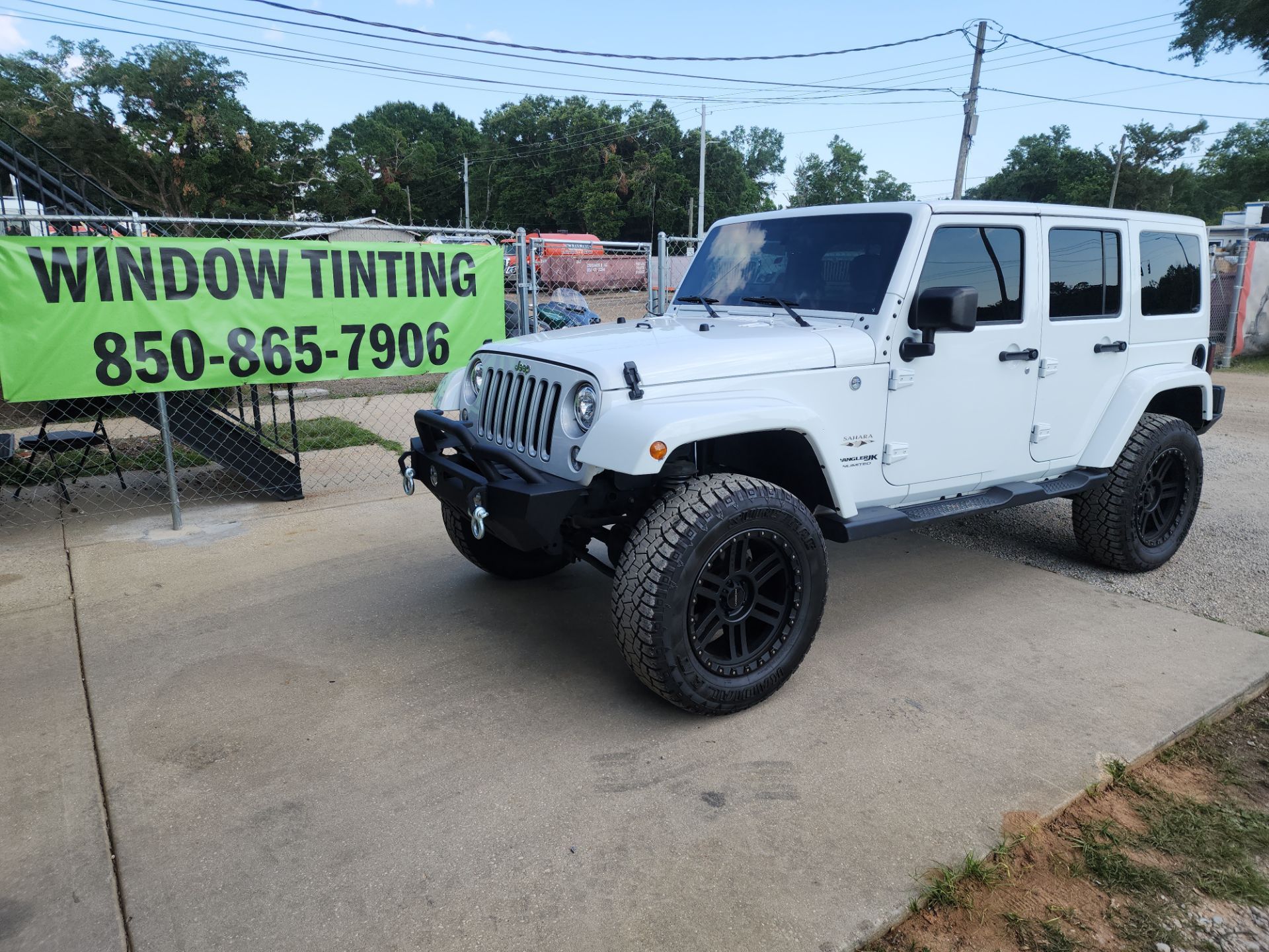 in the photo there is a white jeep wrangler parked in front of a banner advertising window tinting services by t's window tinting the jeep features aftermarket wheels which suggest it may be customized in other ways as well including a potential new window tint job window tinting involves applying a thin laminate film to the interior or exterior of the glass surfaces on a vehicle significantly the windows to reduce the amount of ultraviolet uv and infrared light that passes through the glass this process can protect the vehicle interior from sun damage provide some shatter resistance and increase privacy for passengers as well as enhance the aesthetics of the vehicle