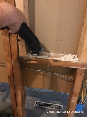A vacuum hose cleaning loose white debris from an exposed wall cavity and wood framing.