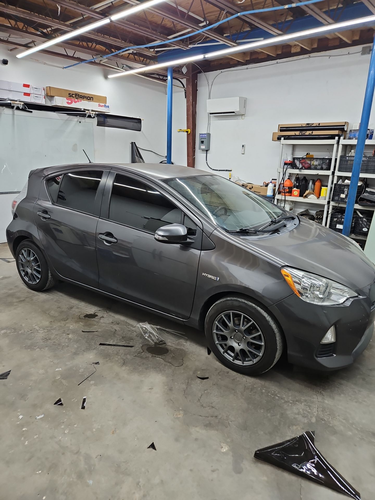 the image shows a car being serviced for window tinting the vehicle is likely a compact hybrid model being worked on in a garage setting the windows are partially tinted giving a sleek appearance there are pieces of window film and other tools scattered on the floor indicating the tinting process is underway the car is probably receiving heat rejection and privacy benefits from the new tint