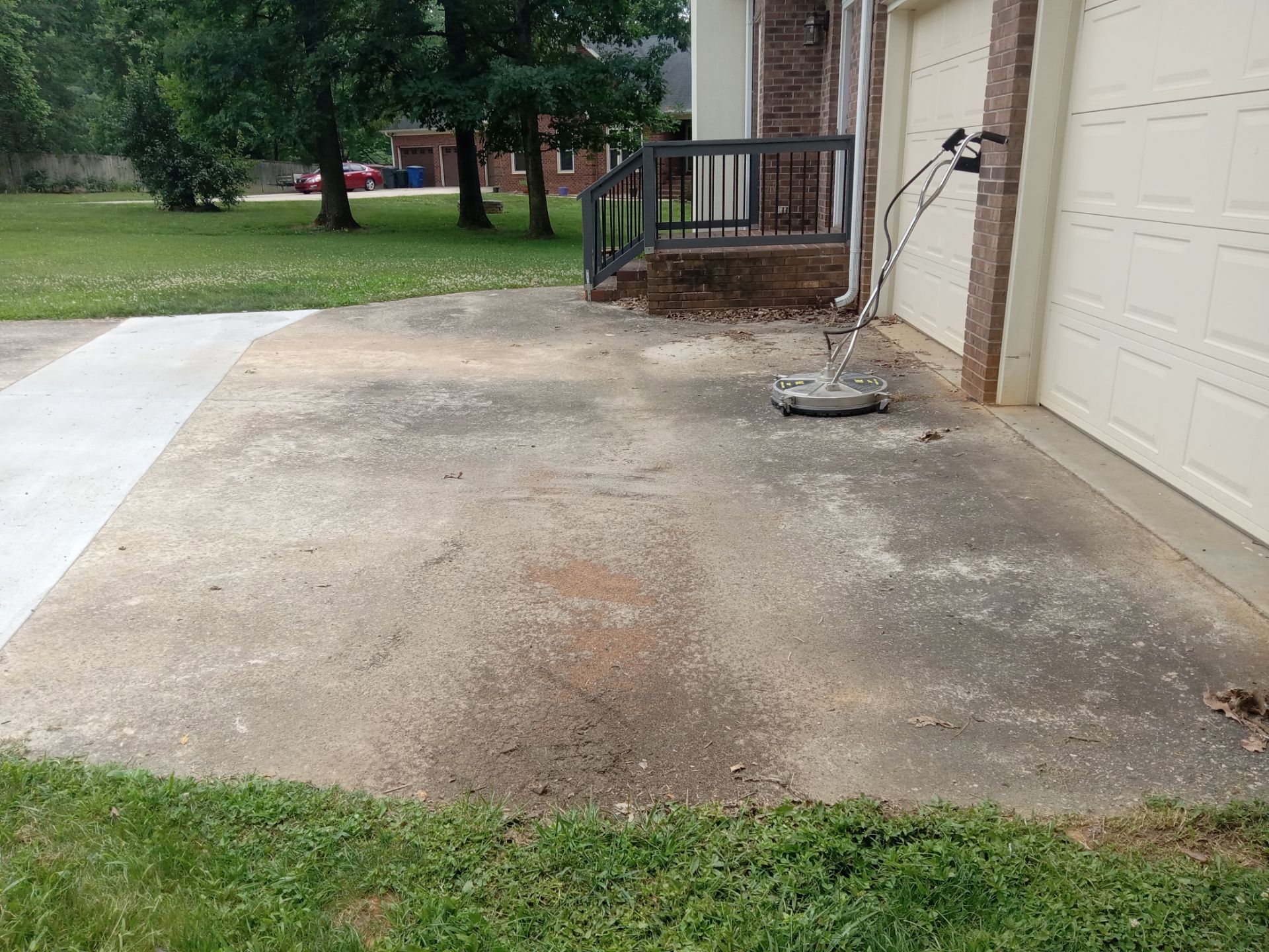 the service being done in the photo involves cleaning a concrete driveway some areas of the driveway appear cleaner indicating that the pressure washing process has begun the concrete surface shows a marked difference between the cleaned and dirty areas a pressure washing tool with a flat circular head is seen which is typically used for evenly and efficiently cleaning flat surfaces such as driveways and sidewalks the surrounding areas include a grassy lawn and part of a residential structure with a brick facade and a garage door