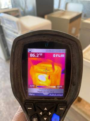 A handheld thermal imaging camera showing a heat map, with boxes and shelving in the background.