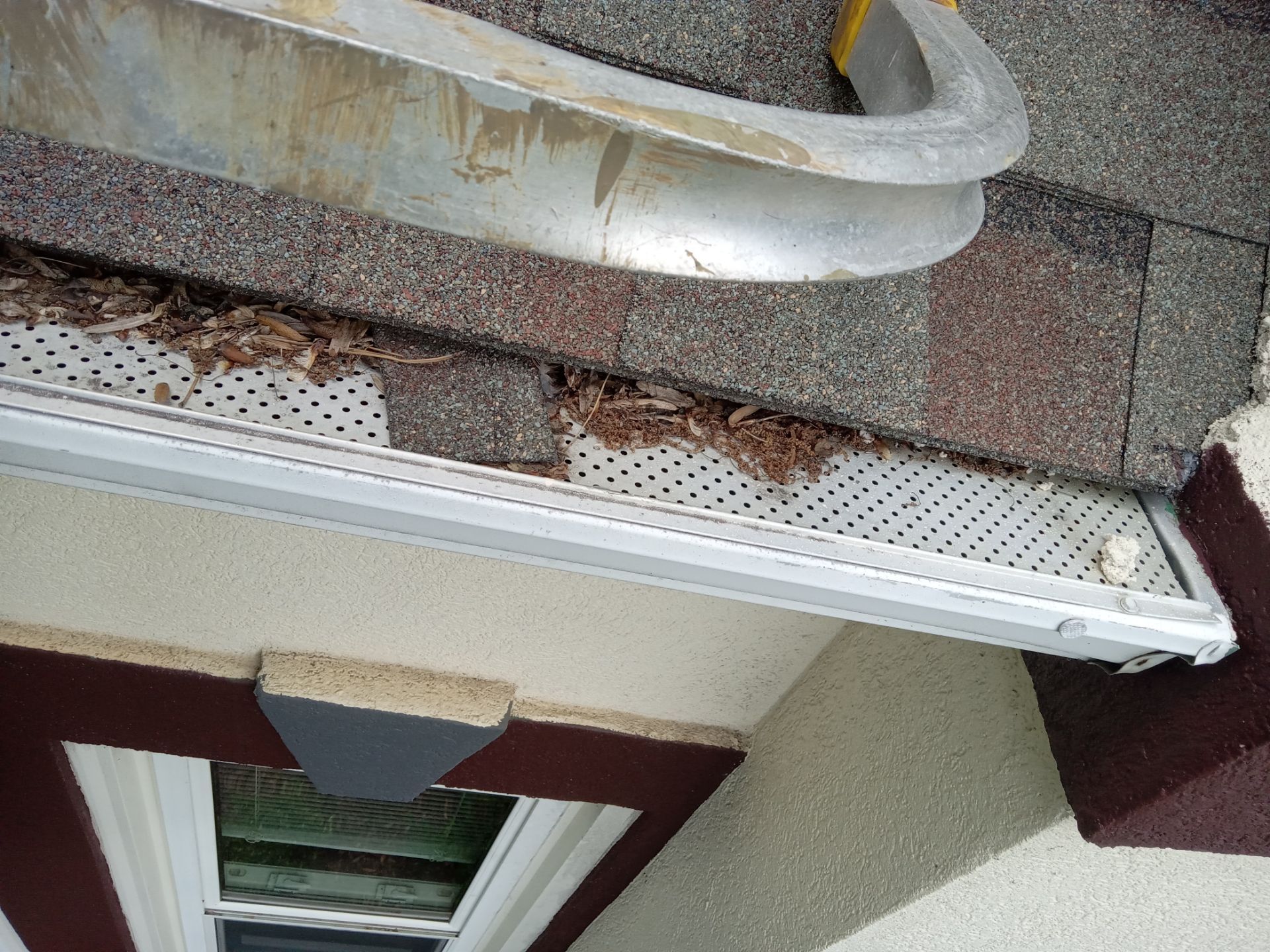 the image shows a close-up view of a house gutter that is clogged with leaves and debris the gutter is installed just below the roof shingles where it is intended to collect rainwater and direct it away from the house exterior and foundation the pressure washing company is likely providing gutter cleaning services to remove the accumulated organic material and ensure proper water flow through the gutters the surface shown in the photo indicates that the cleaning has not yet been performed or is in the process of being done