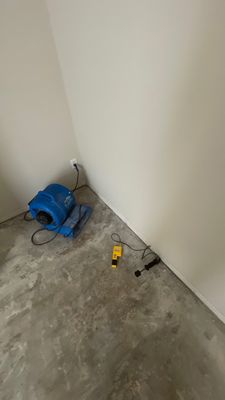 Blue air mover and yellow moisture meter placed on a concrete floor near a wall corner.