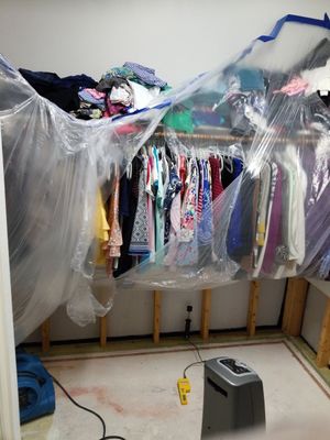 Closet with hanging clothes covered by plastic sheeting, exposed lower wall studs, and drying equipment on the floor.