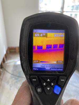 A handheld FLIR thermal imaging camera displaying a wall and floor scan inside a commercial room.