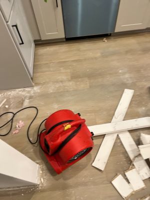 A red air mover on a kitchen floor near removed trim pieces and small debris.