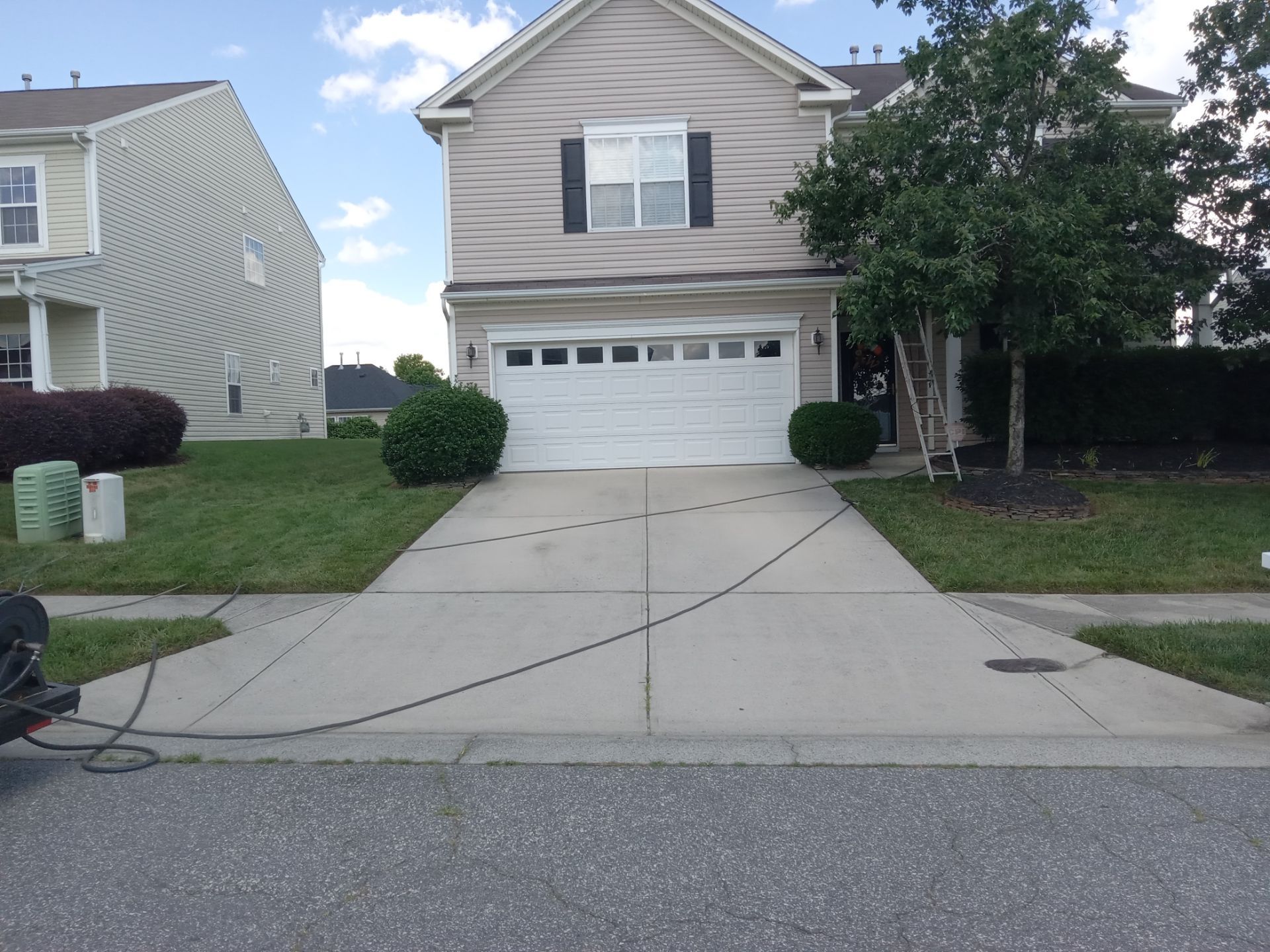 in the photo a pressure washing service is cleaning the concrete driveway the before and after difference is shown by a clean stripe down the middle of the driveway where the pressure washing has removed dirt and stains the house front also appears to be in line for cleaning given the presence of a ladder leaning against the house indicating the cleaning service may include exterior house washing as well as driveway cleaning