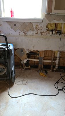 Kitchen wall with removed lower drywall, exposed plumbing, cords, and drying equipment nearby.