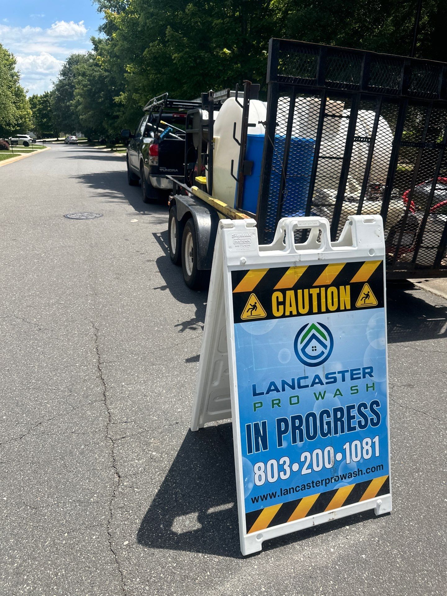 in the image there is a caution sign indicating a pressure washing service by lancaster pro wash the sign is placed on a paved road suggesting the pressure washing may involve cleaning concrete surfaces such as driveways sidewalks or parking lots the presence of a vehicle with a mounted tank and equipment on a trailer behind it implies that the company is equipped to handle large-scale or commercial pressure washing tasks the service likely includes the removal of dirt grime mold or other buildup from outdoor surfaces using high-pressure water jets