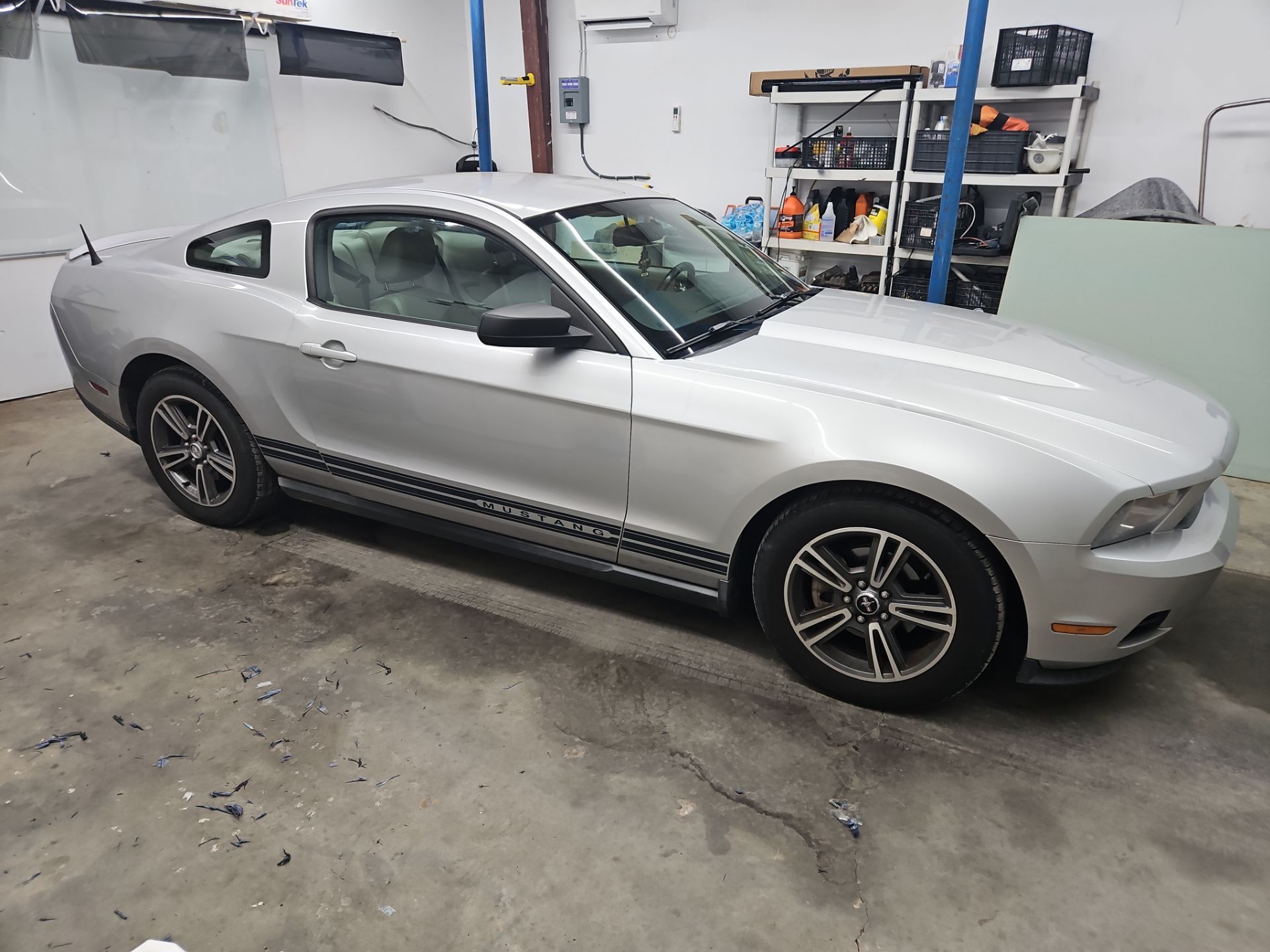 a silver mustang is having its windows tinted by t's window tinting in pensacola fl the car is inside a garage or workshop setting there are various tools and supplies on shelves nearby the tint film is likely being applied to all windows of the car to reduce heat glare and uv exposure while providing a sleek look