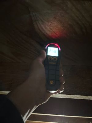 A handheld moisture meter being used against a wood surface in a dim crawlspace area.