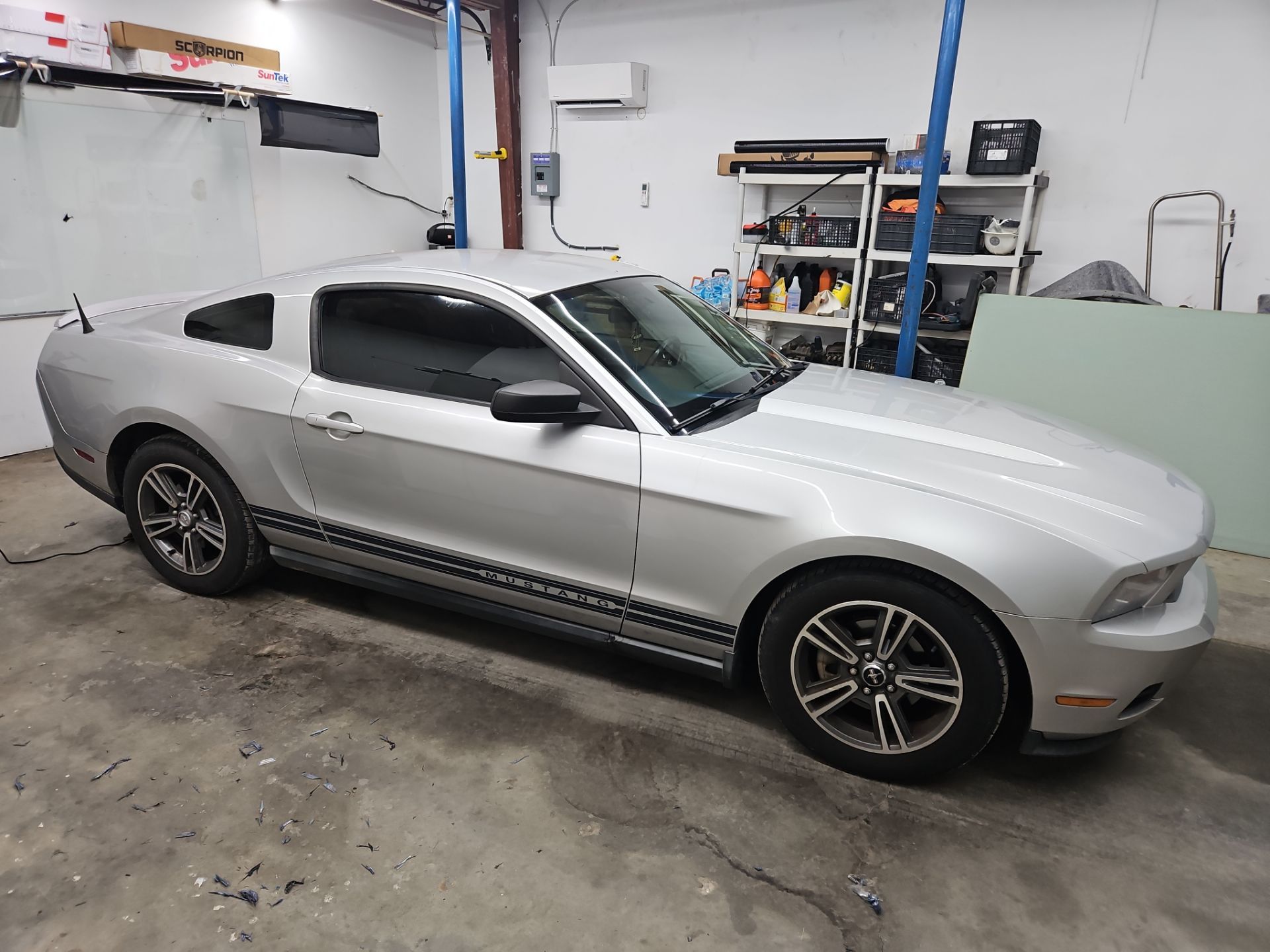 window tinting service is being done on a silver sports car the car is a ford mustang and appears to be in a workshop the windows have been tinted dark likely to reduce glare and provide privacy the floors have small pieces of window film suggesting ongoing tinting work the car is in the process of having tint applied or has just completed the tinting service