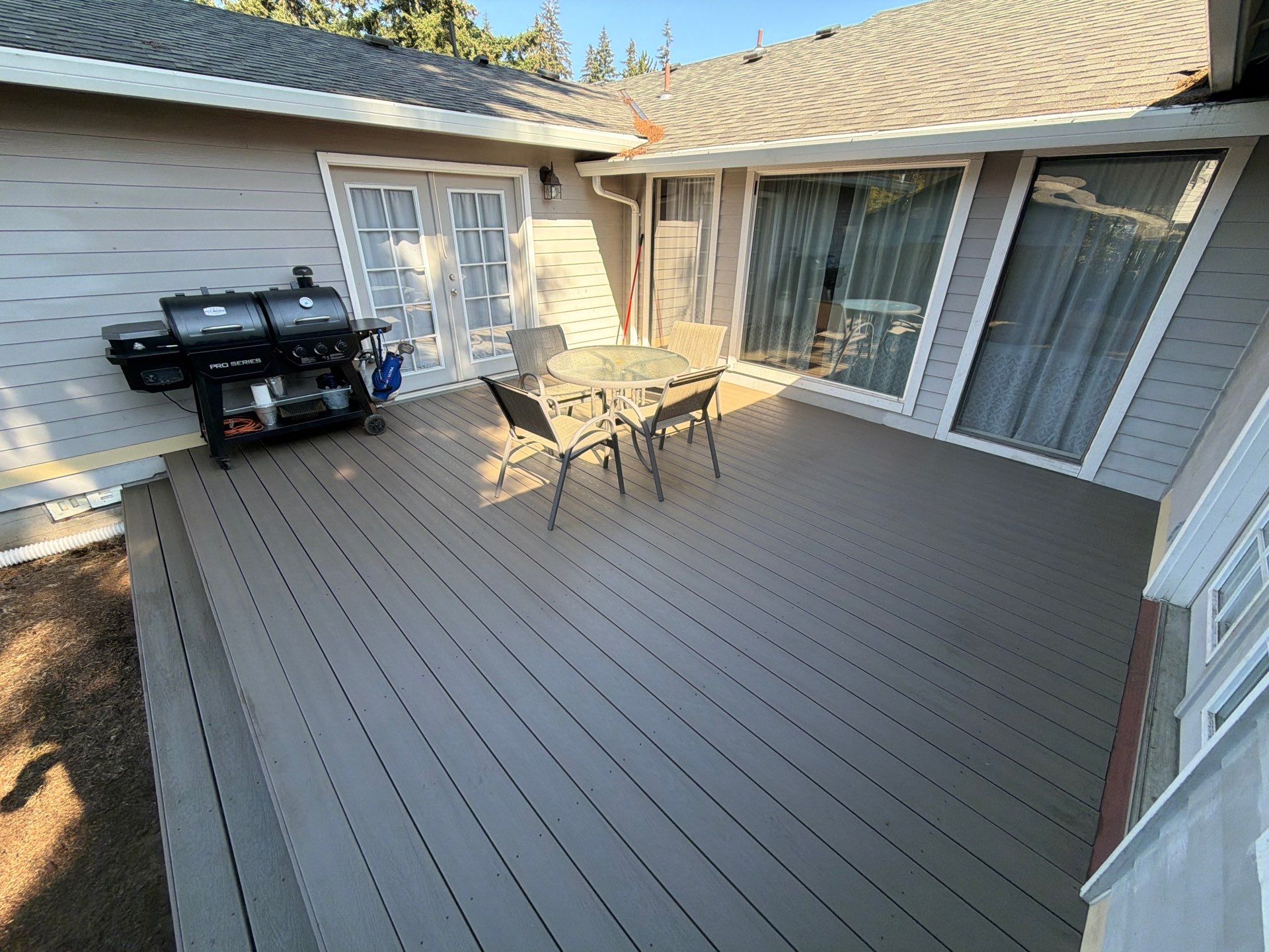 A composite deck area extends from the rear of a single-story residence, featuring a black gas grill positioned against light gray lap siding near white French doors. A small circular glass-top patio table surrounded by four matching chairs sits in the center of the deck, while large glass sliding doors and windows provide views into the home interior. To the left of the deck, a patch of exposed dirt is visible, with mature trees partially visible above the asphalt shingle roofline in the background.