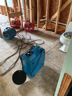 A partially demolished bathroom with exposed wall studs, subfloor, toilet, cords, fans, and a blue dehumidifier.
