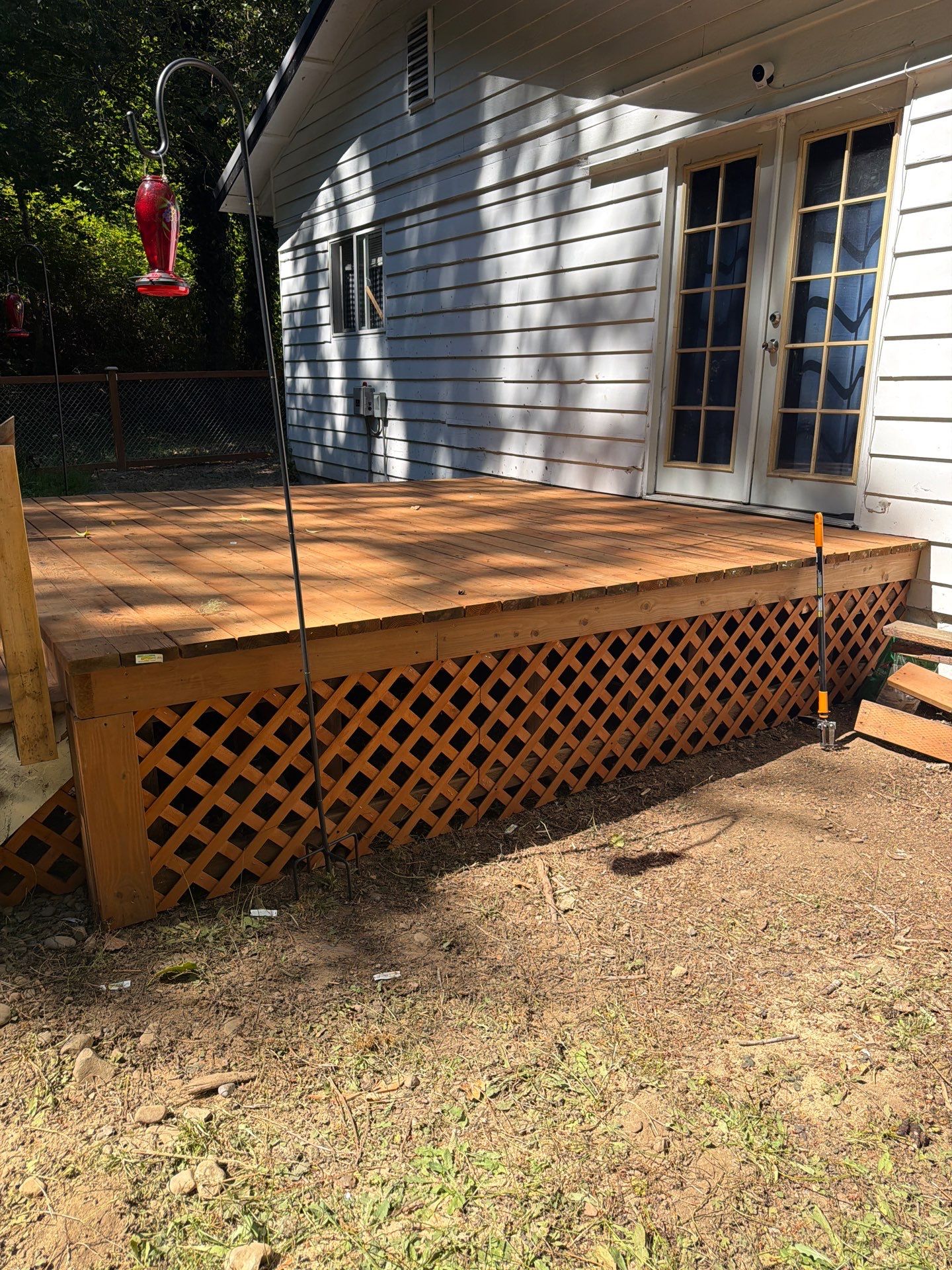 A newly constructed wooden deck with lattice skirting is attached to the rear of a white-sided house featuring French doors. A black metal bird feeder stand is positioned in the foreground near the deck, while the surrounding yard consists of bare dirt and sparse vegetation.