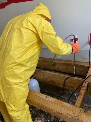 A remediation worker in a yellow protective suit applying treatment near exposed floor framing.