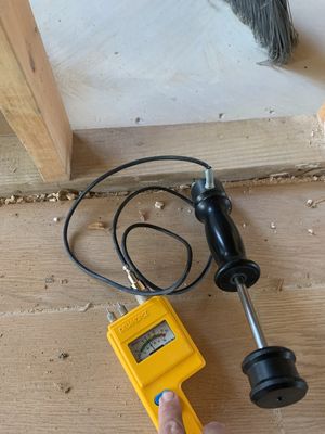A moisture meter probe being used near the base of an exposed wall and subfloor.