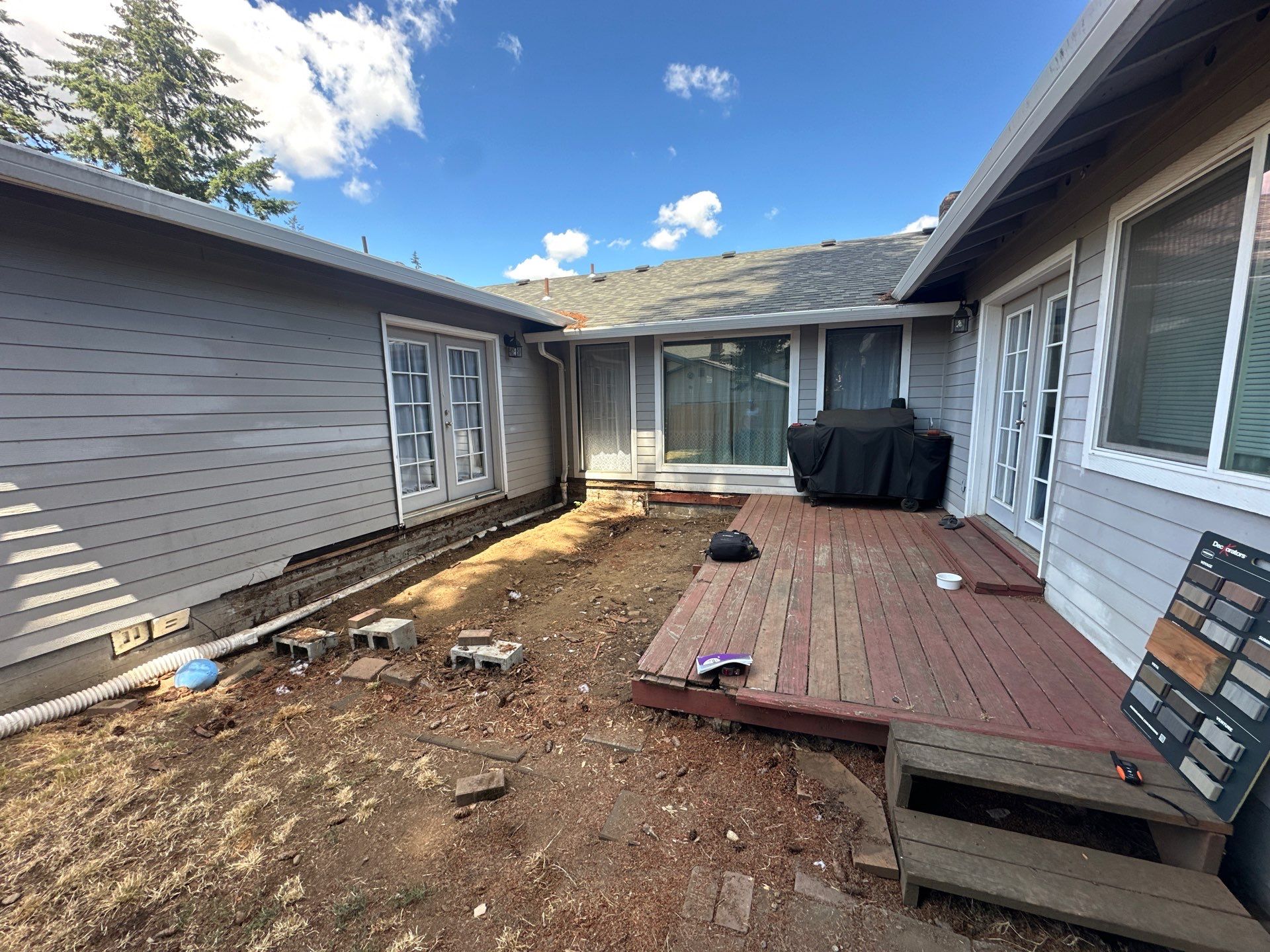 A backyard area features a wooden deck adjacent to a grey-sided house with white-framed French doors and windows. The ground surrounding the deck consists of exposed dirt and scattered construction debris including concrete blocks, while a black grill sits on the deck surface near a sample display board leaning against the house exterior wall.