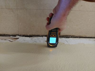 A hand holding a moisture meter against the lower wall near the floor.