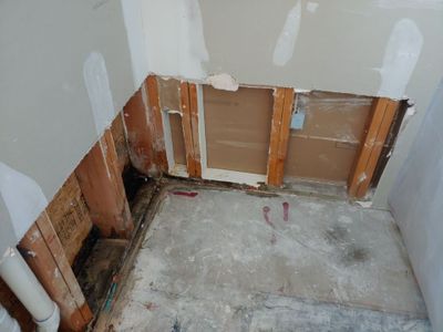 Small room with lower drywall removed, exposed wood framing, concrete slab floor, and plumbing along one wall.