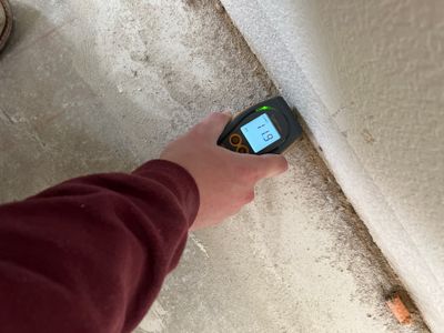 Moisture meter held against a concrete floor beside a wall and baseboard area.