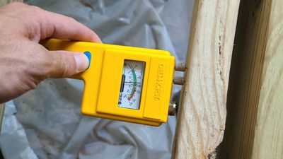 A hand holding a yellow moisture meter against a wood framing member.