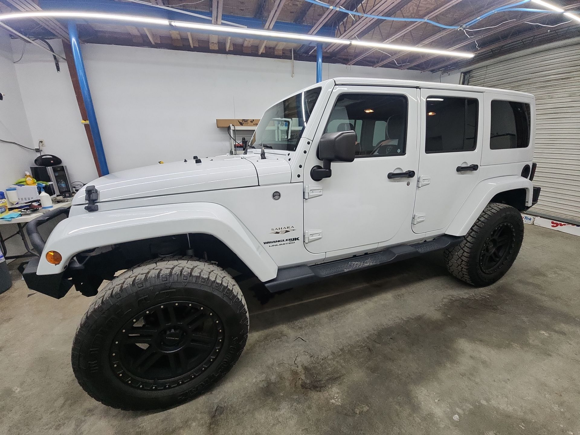 the image shows a white jeep wrangler in a garage likely undergoing window tinting by the company mentioned the jeep has multiple doors and windows which will have a film applied to them for reducing glare and heat as well as providing privacy the process involves cleaning the windows cutting and shaping the tint material and applying it carefully to avoid bubbles and imperfections the surroundings suggest this service is being done indoors to minimize dust and debris during application