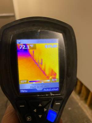 A handheld FLIR thermal imaging camera displaying a temperature reading and wall surface scan.